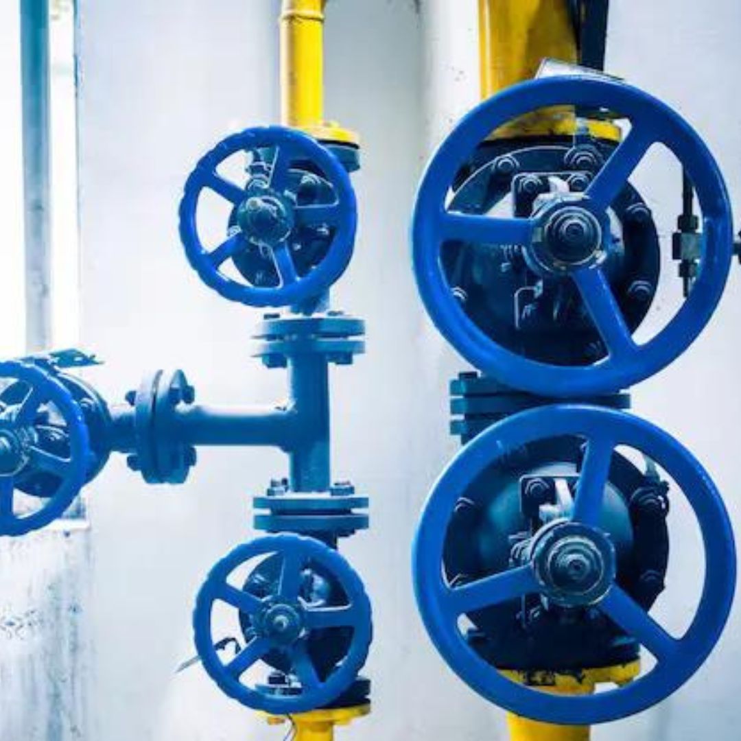 A close-up view of four blue industrial valves with wheel handles, attached to pipes against a white and yellow background.